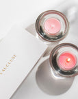 An overhead top-down view of two burning pink rose tealight candles in clear glass holders next to a white Vaucluse luxury gift box.
