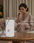 A premium Vaucluse lychee reed diffuser and box set displayed on a wooden table, with a woman in a beige robe relaxing on a couch in a softly lit living room.