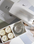 Luxury vanilla scented tealights and clear glass holder arranged in an open branded white box over chic editorial design books.