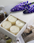 A premium white Vaucluse 16-pack box sliding open to reveal the top tier of cream musk tealight candles, elegantly styled under a cluster of vibrant purple flowers.