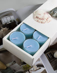 A premium white Vaucluse 16-pack box sliding open to reveal a tier of light blue sea breeze tealight candles, decorated with a textured white seashell.