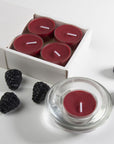 An overhead top-down view of two deep purple blackberry tealight candles in modern clear glass holders resting next to a white embossed Vaucluse gift box.