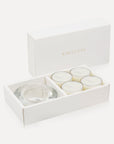An open luxury white gift box displaying a set of four creamy white jasmine tealight candles next to a round clear glass candle holder.