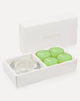 An open luxury white gift box cleanly displaying a set of four vibrant green lily tealight candles alongside a thick round clear glass candle holder.