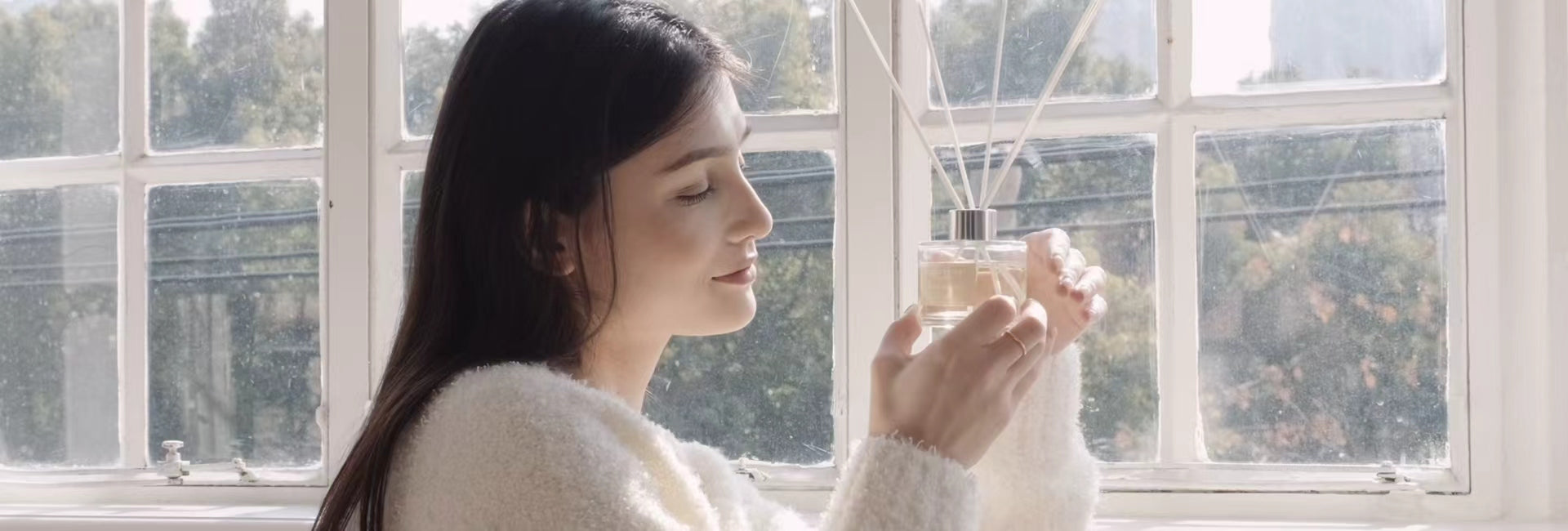 A woman in a cozy sweater holding a Vaucluse Fragrance luxury reed diffuser by a bright window, enjoying the home fragrance.