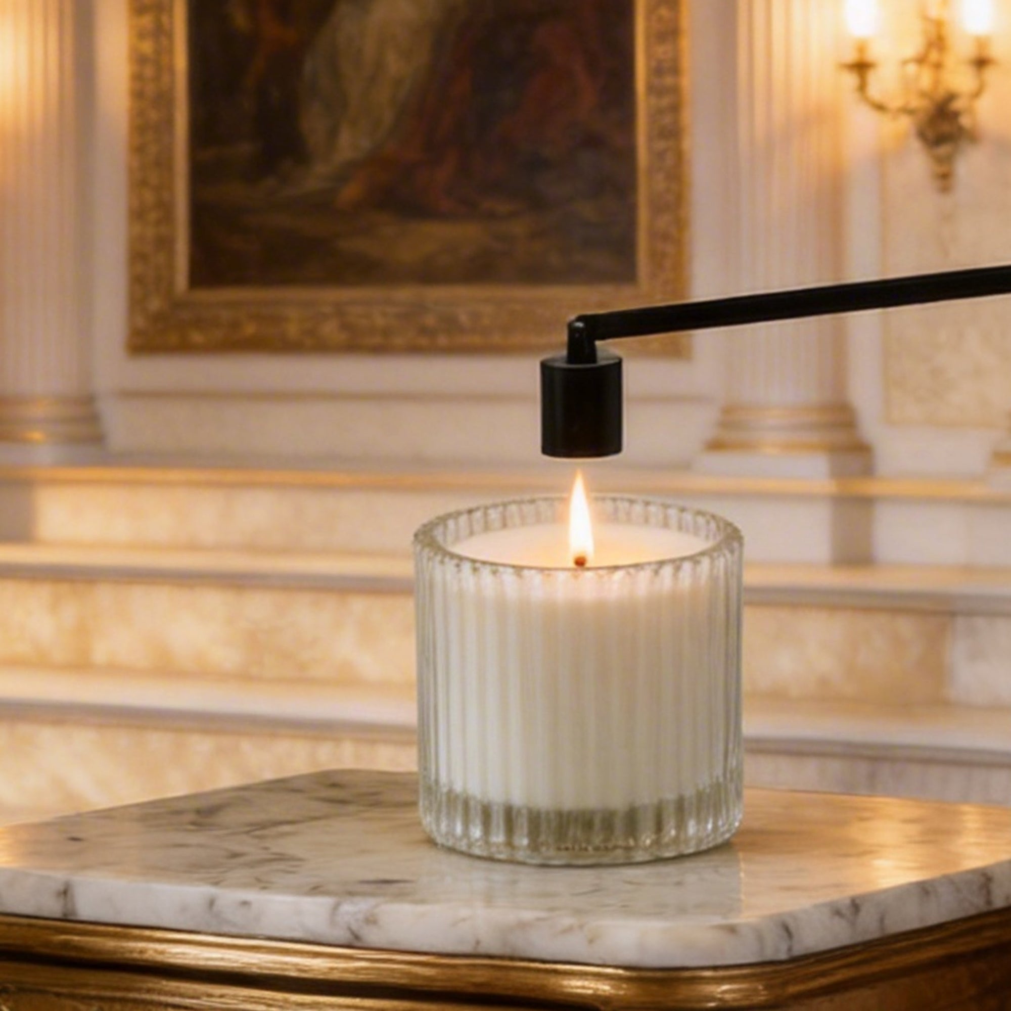 A matte black bell candle snuffer being used to safely extinguish the flame of a luxury white candle on a marble table.