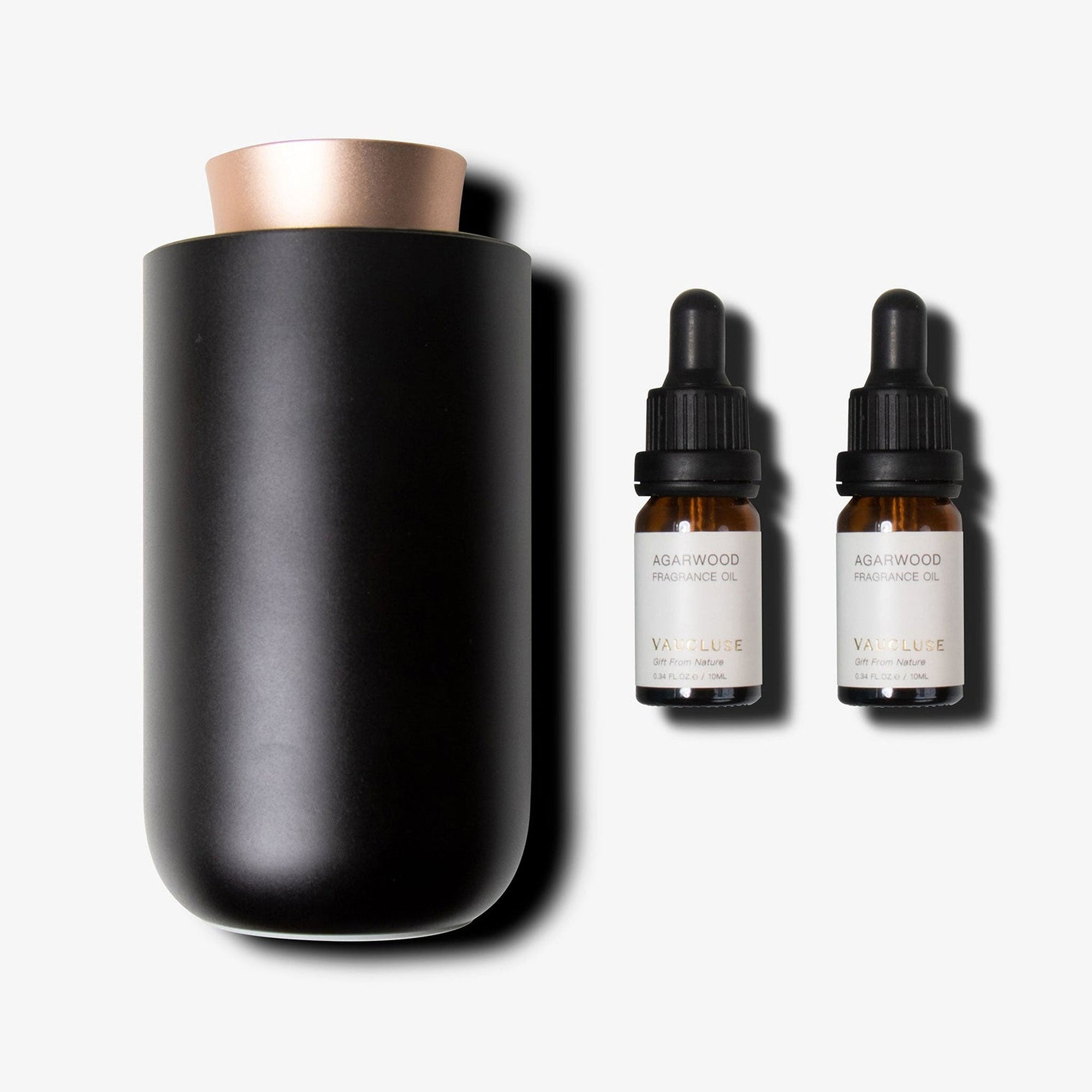 A matte black waterless aromatherapy diffuser with a rose gold accent lid standing next to two amber glass essential oil dropper bottles.