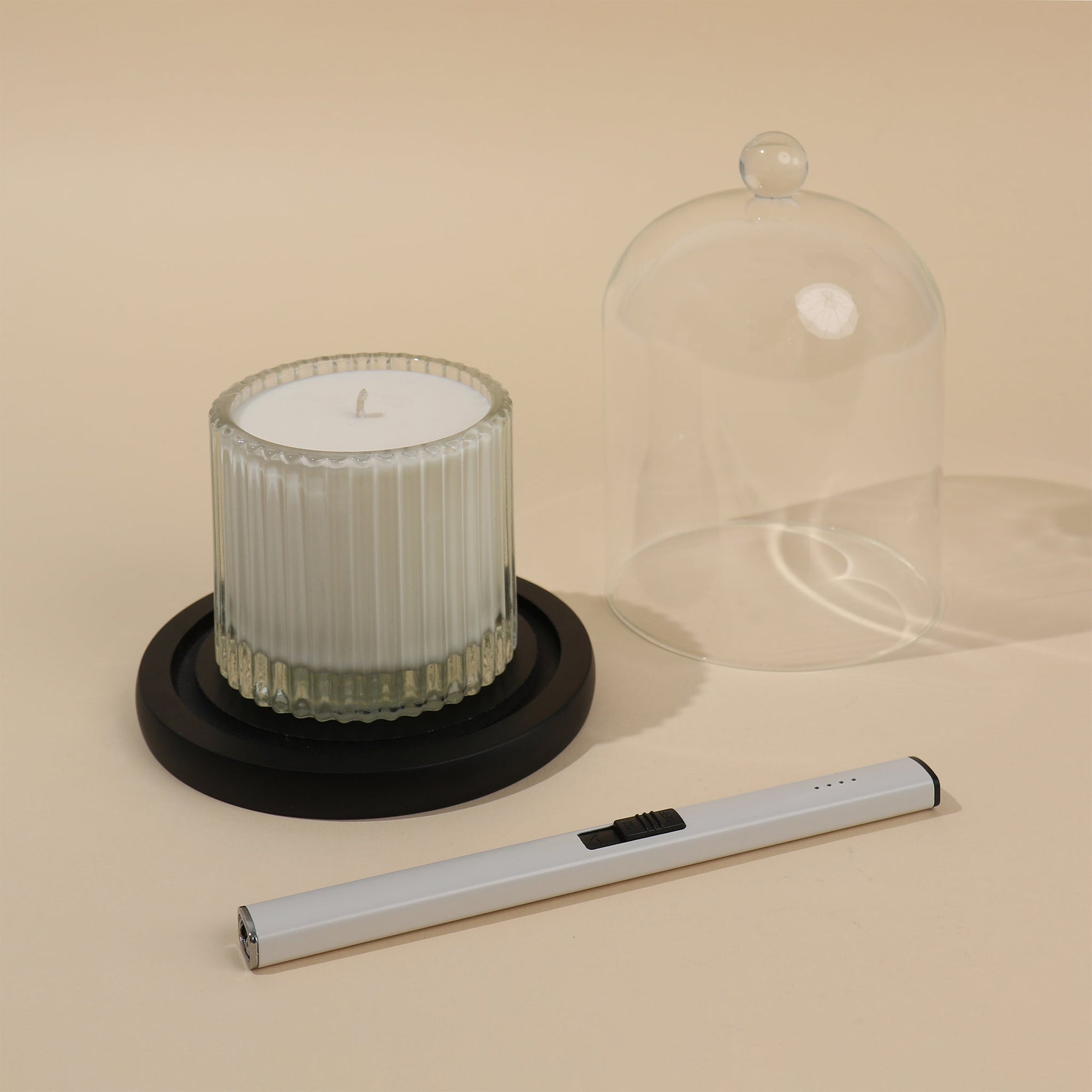 A luxury glass candle dome removed from its black base to reveal a white ribbed jar candle next to a sleek electric lighter on a beige surface.