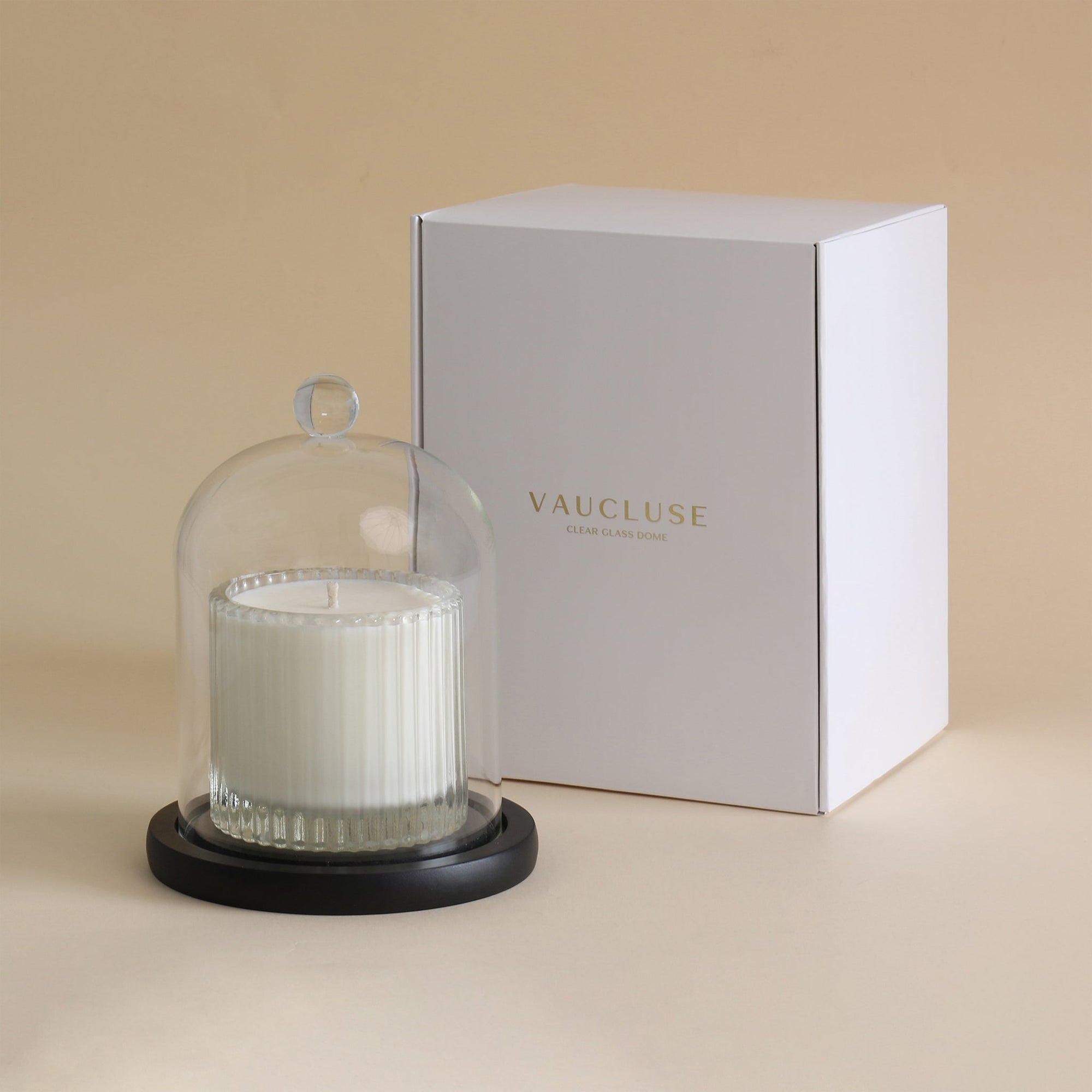 The clear glass cloche dome housing a white jar candle on its black base, positioned in front of an elegant white Vaucluse branded gift packaging box.