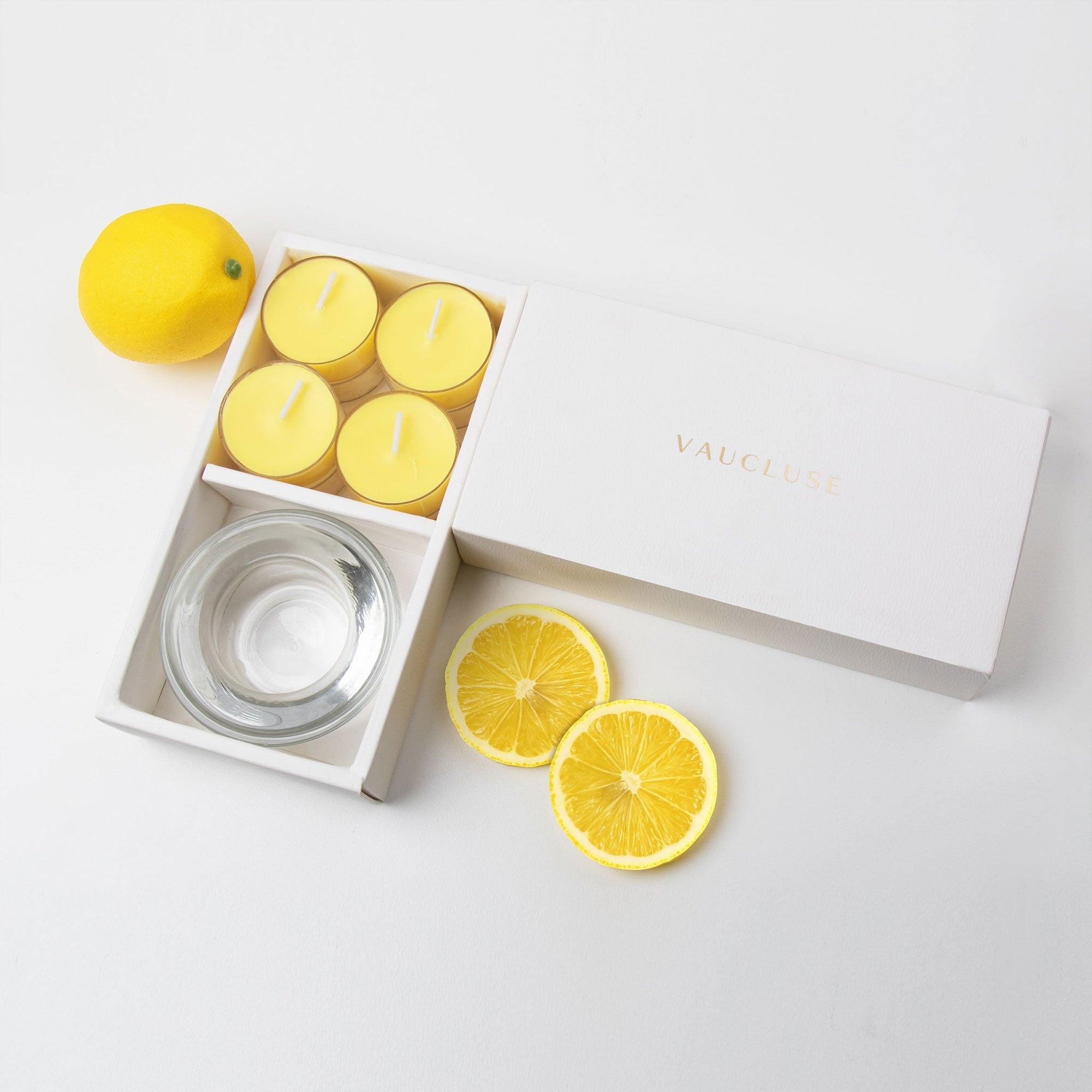 Minimalist overhead flat lay of the open yellow tealight candle set styled with a whole fresh lemon and two vibrant citrus slices on a white background.