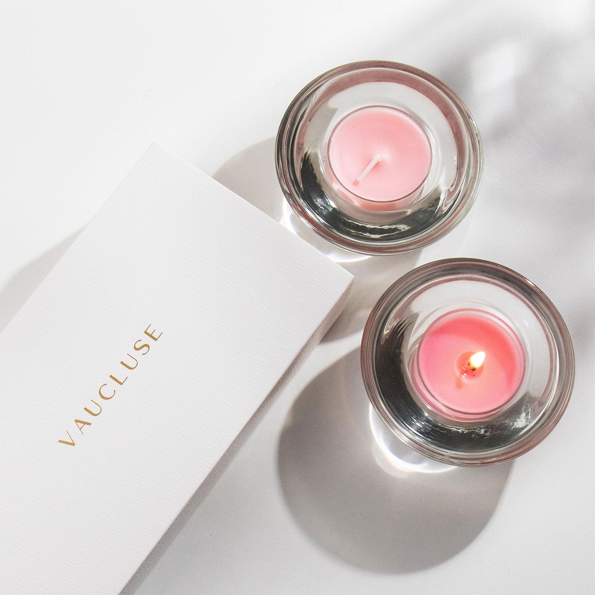 An overhead top-down view of two burning pink rose tealight candles in clear glass holders next to a white Vaucluse luxury gift box.