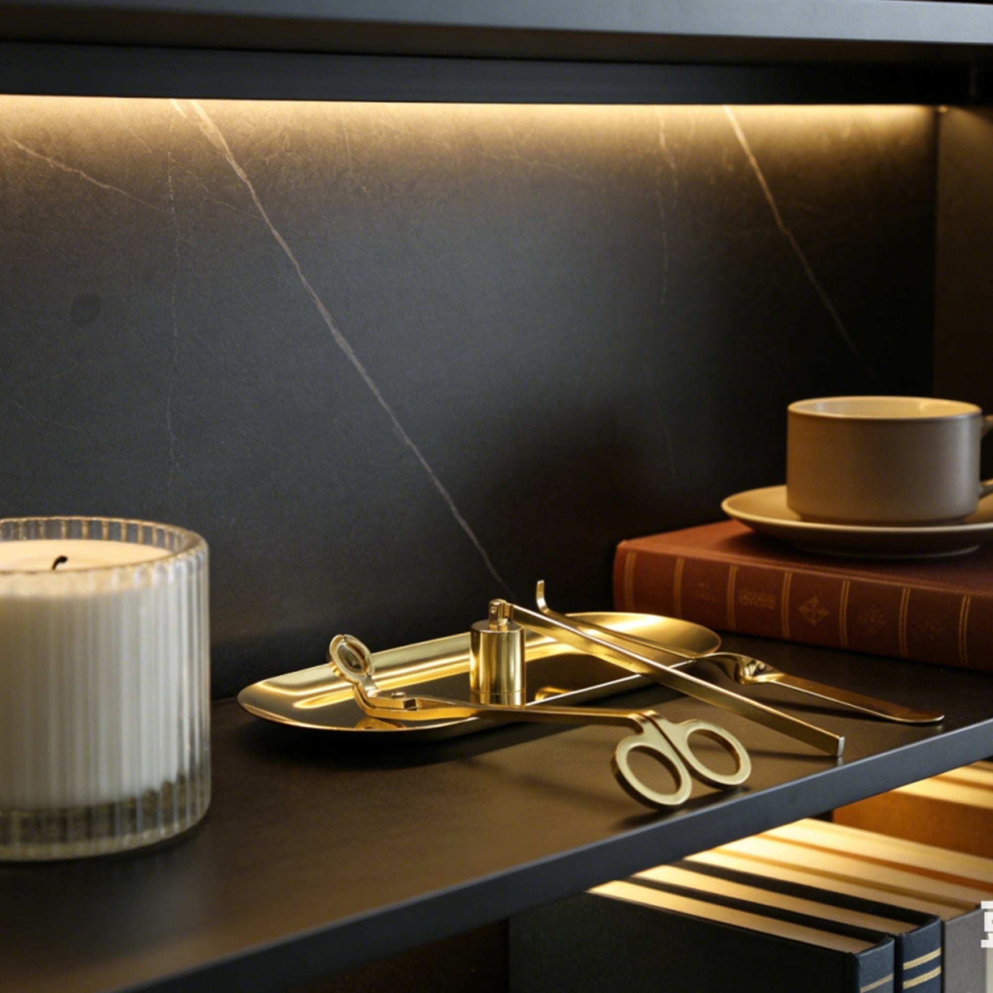 An elegant gold candle care set elegantly arranged on its matching tray resting on a dark modern bookshelf next to a ribbed candle.