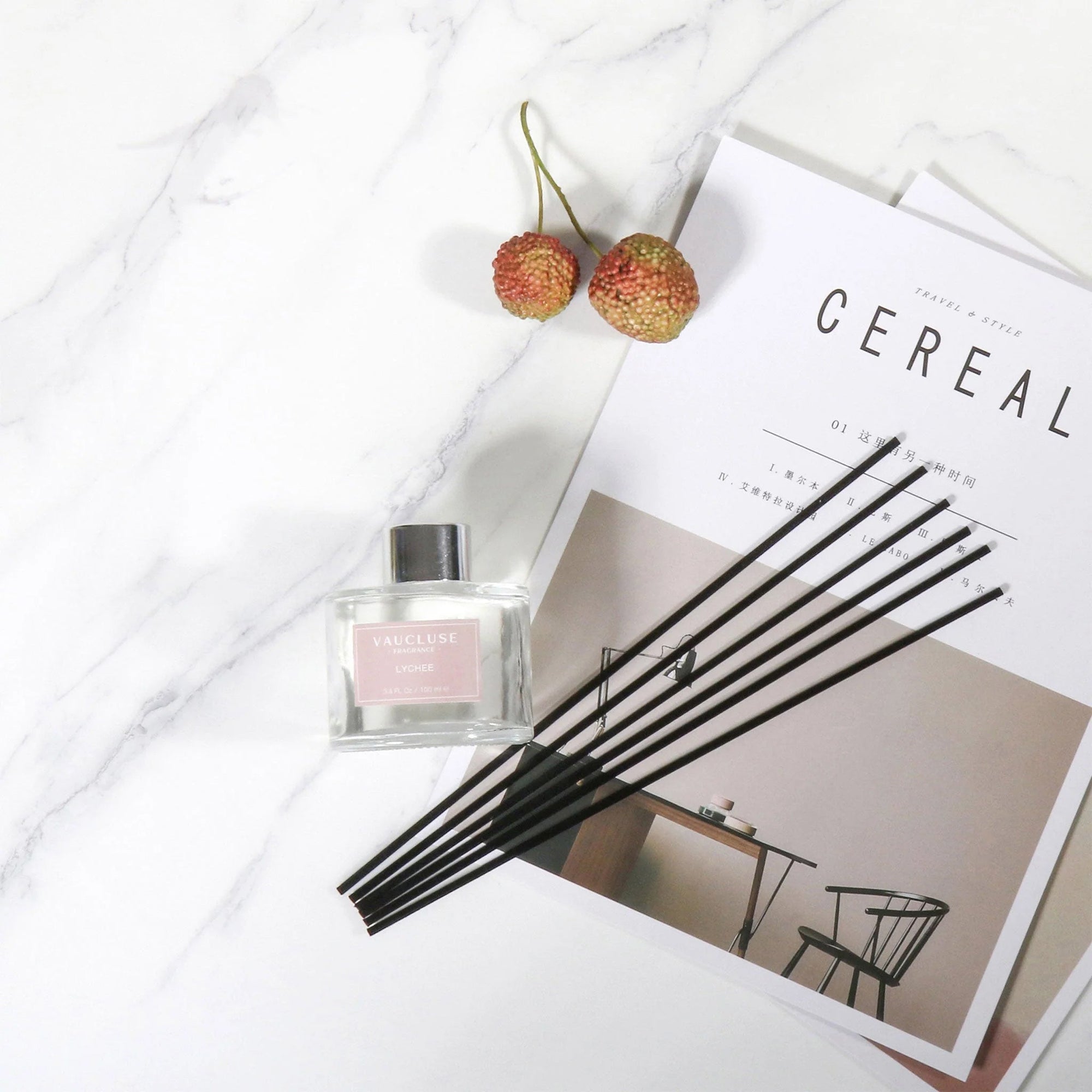 An overhead flat lay view of a clear glass Vaucluse diffuser bottle and its black reeds arranged on an aesthetic magazine alongside fresh lychee fruits on a marble surface.