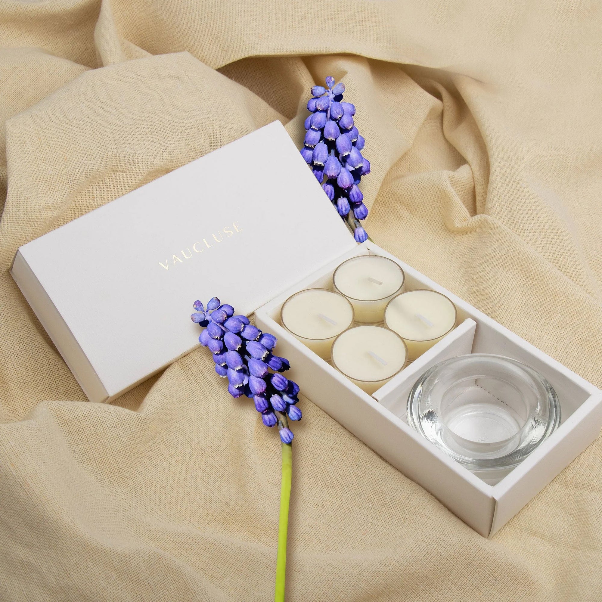 Elegant aromatherapy lifestyle scene featuring the white musk tealight gift box styled on draped beige linen fabric with purple floral accents.