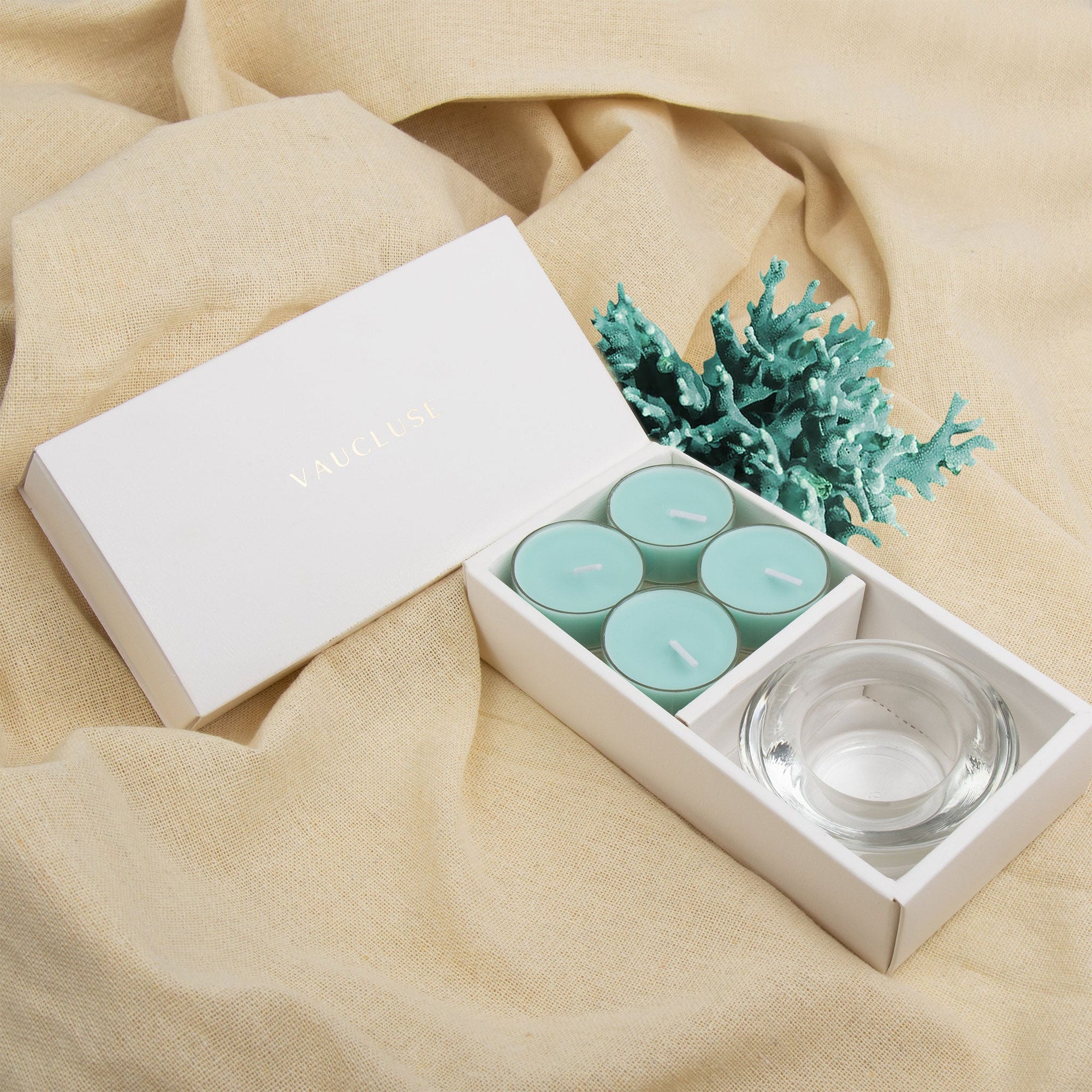 Relaxing lifestyle scene showing the light blue ocean tealight set and clear glass holder resting on textured beige linen fabric next to sea coral decor.
