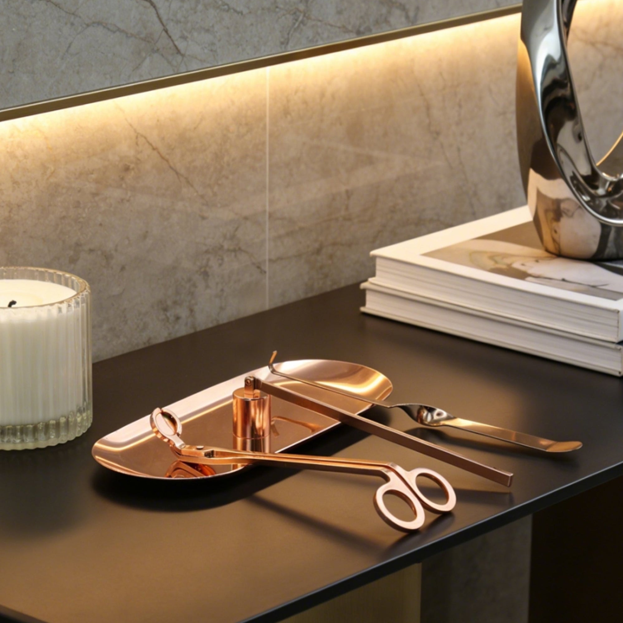An elegant rose gold candle care set arranged on its matching tray resting on a sleek dark modern desk.
