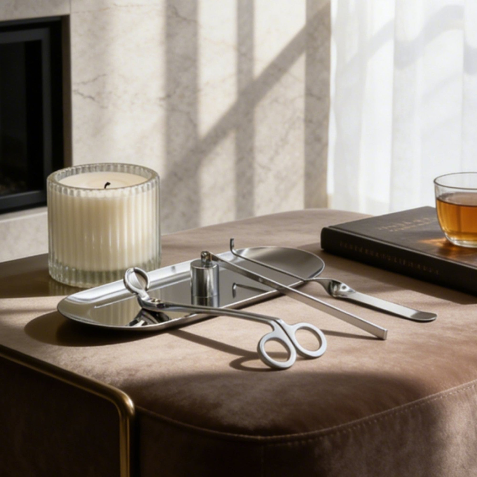An elegant silver candle care set arranged on its matching tray resting on a plush brown velvet ottoman next to a ribbed candle.