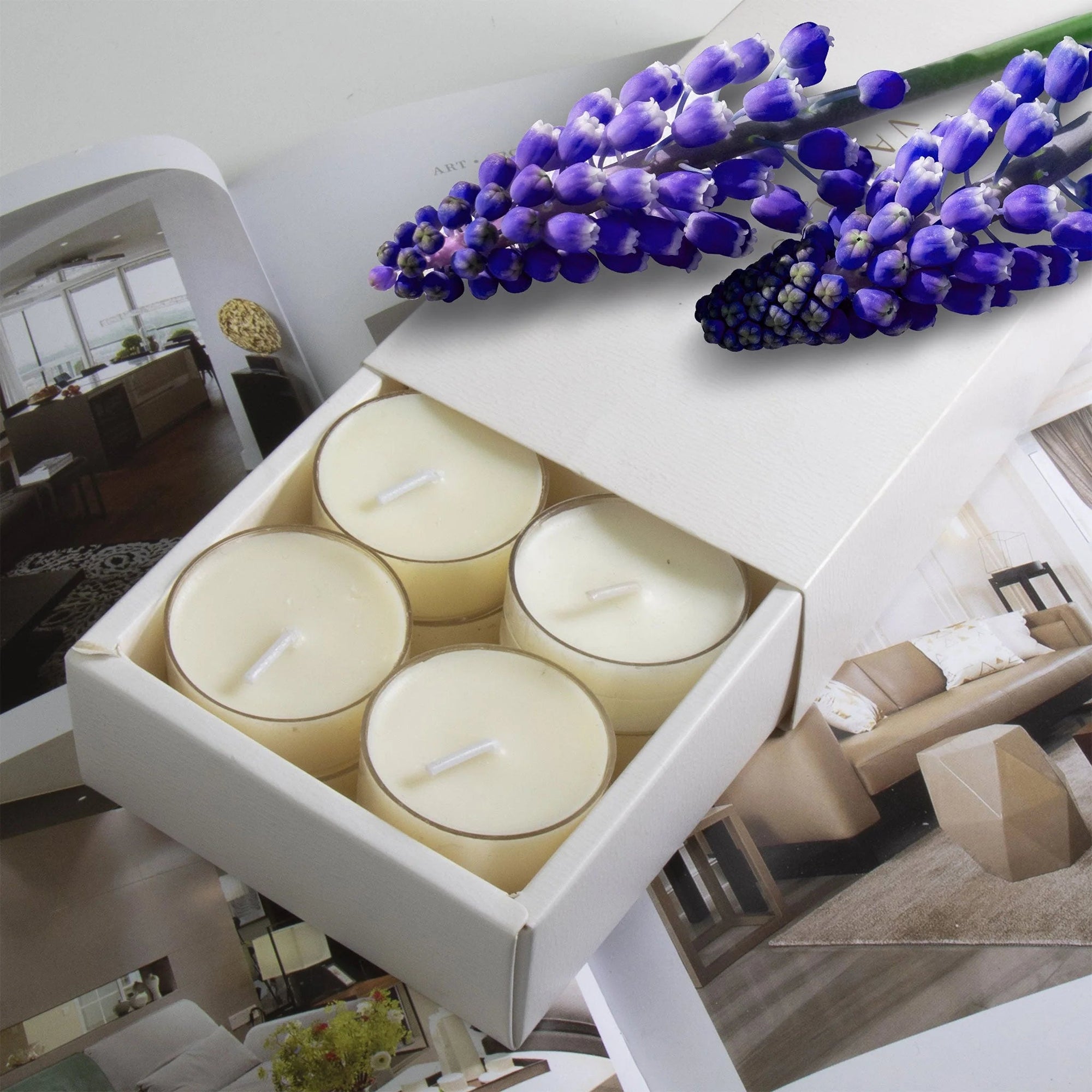 A premium white Vaucluse 16-pack box sliding open to reveal the top tier of cream musk tealight candles, elegantly styled under a cluster of vibrant purple flowers.