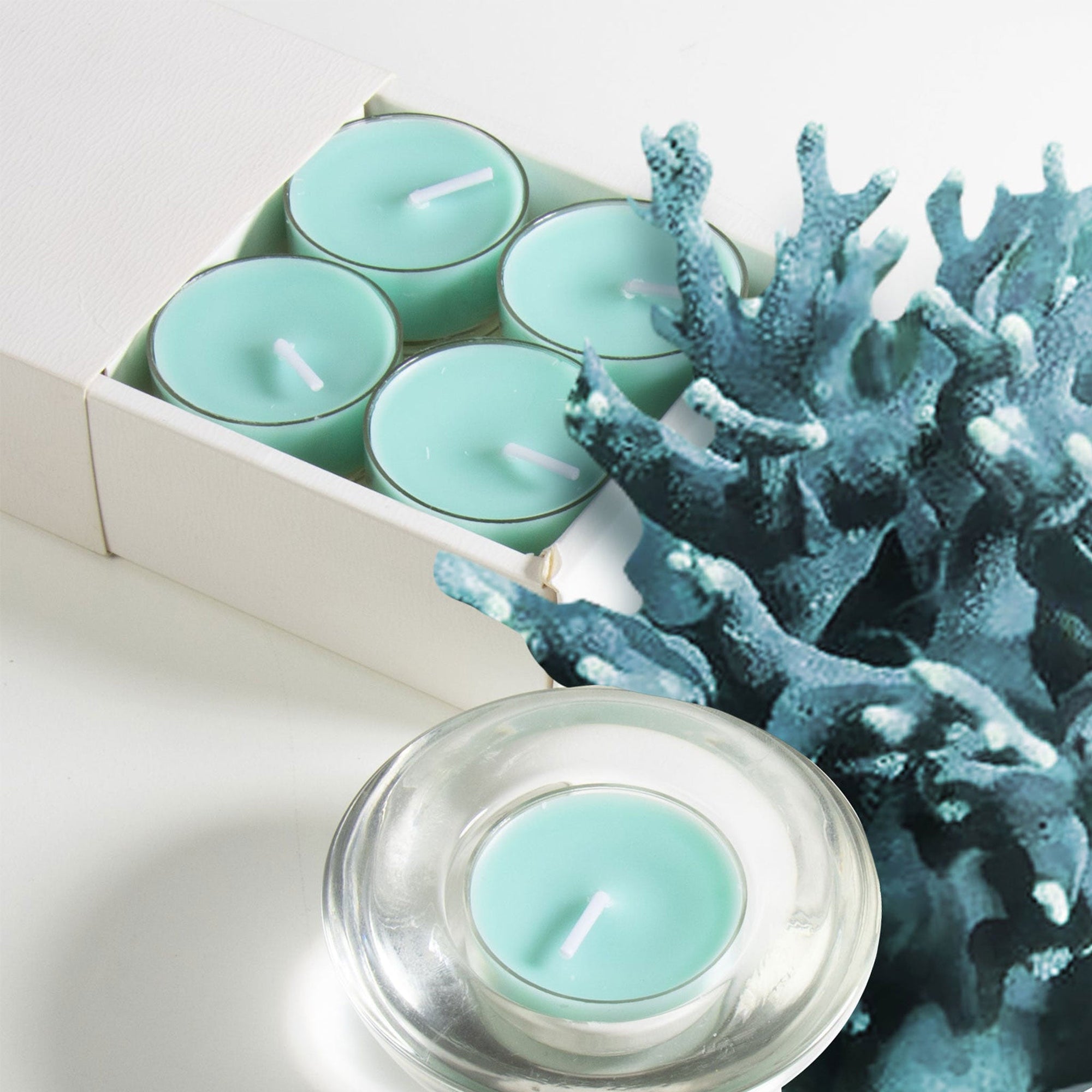 A partial view of light teal ocean scented tealight candles in a box next to a piece of decorative dark blue sea coral and a candle in a clear glass holder