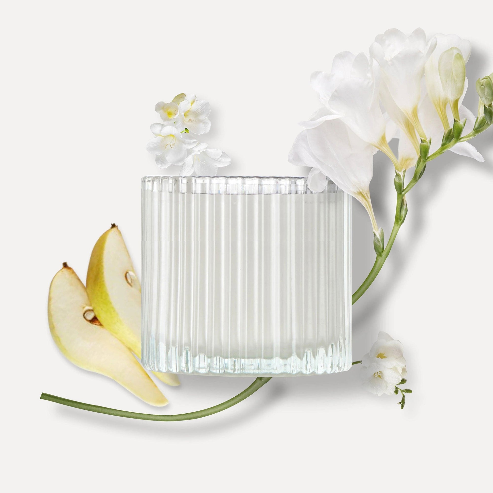 Vaucluse ribbed glass candle jar surrounded by fresh sliced pears and white freesia flowers against a clean background, representing the product's fragrance notes.