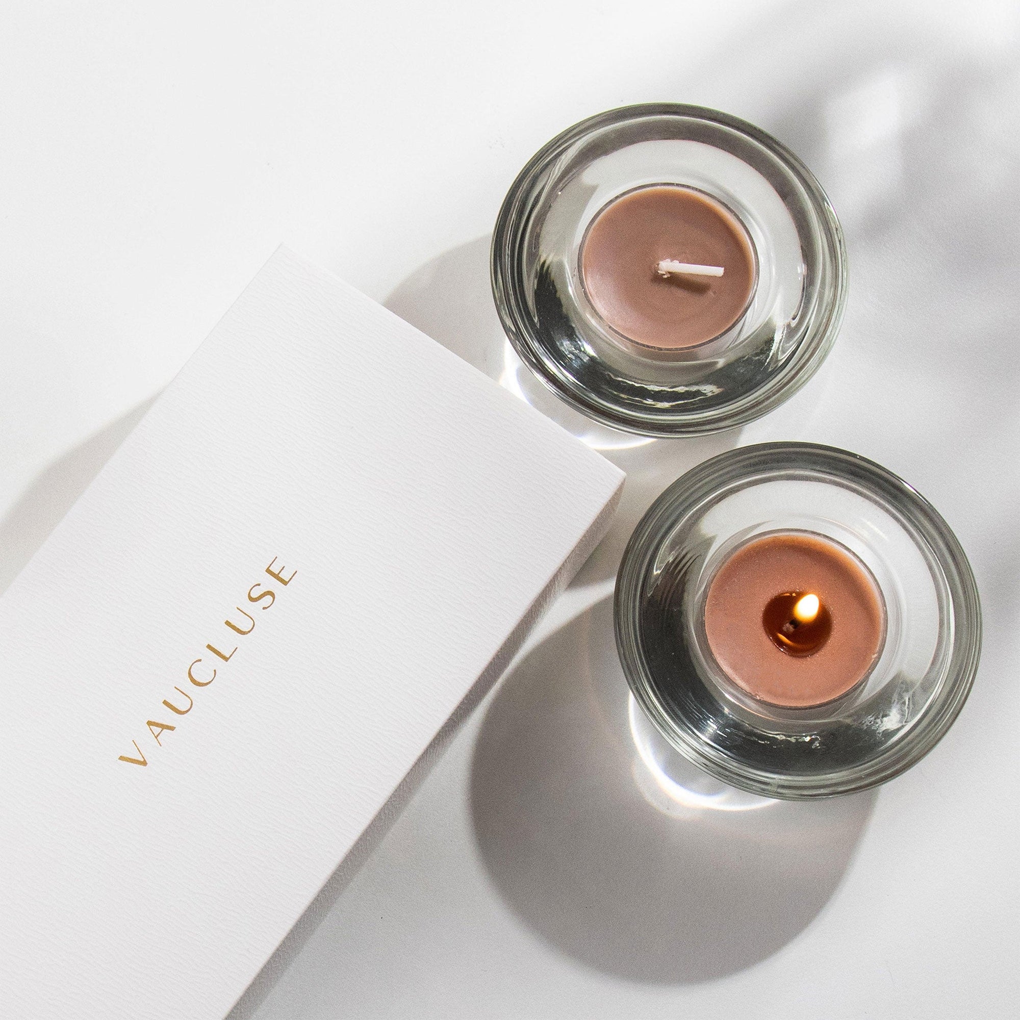 Overhead view of two brown Vaucluse Cinnamon tealight candles in circular glass holders, with one lit and casting a warm glow next to white brand packaging.