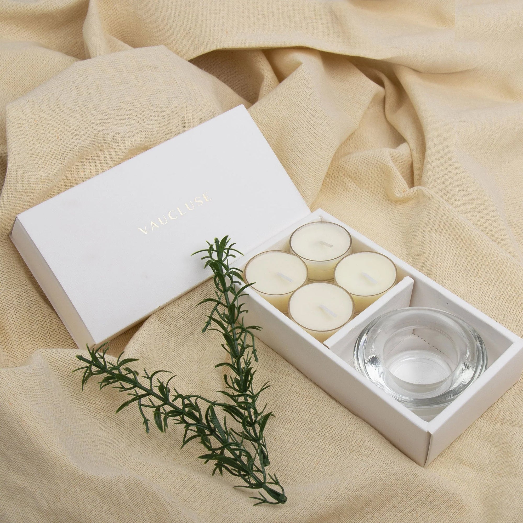 Premium Vaucluse vanilla tealight candles and heavy glass holder set nestled on an aesthetic, softly draped neutral linen background with rosemary.