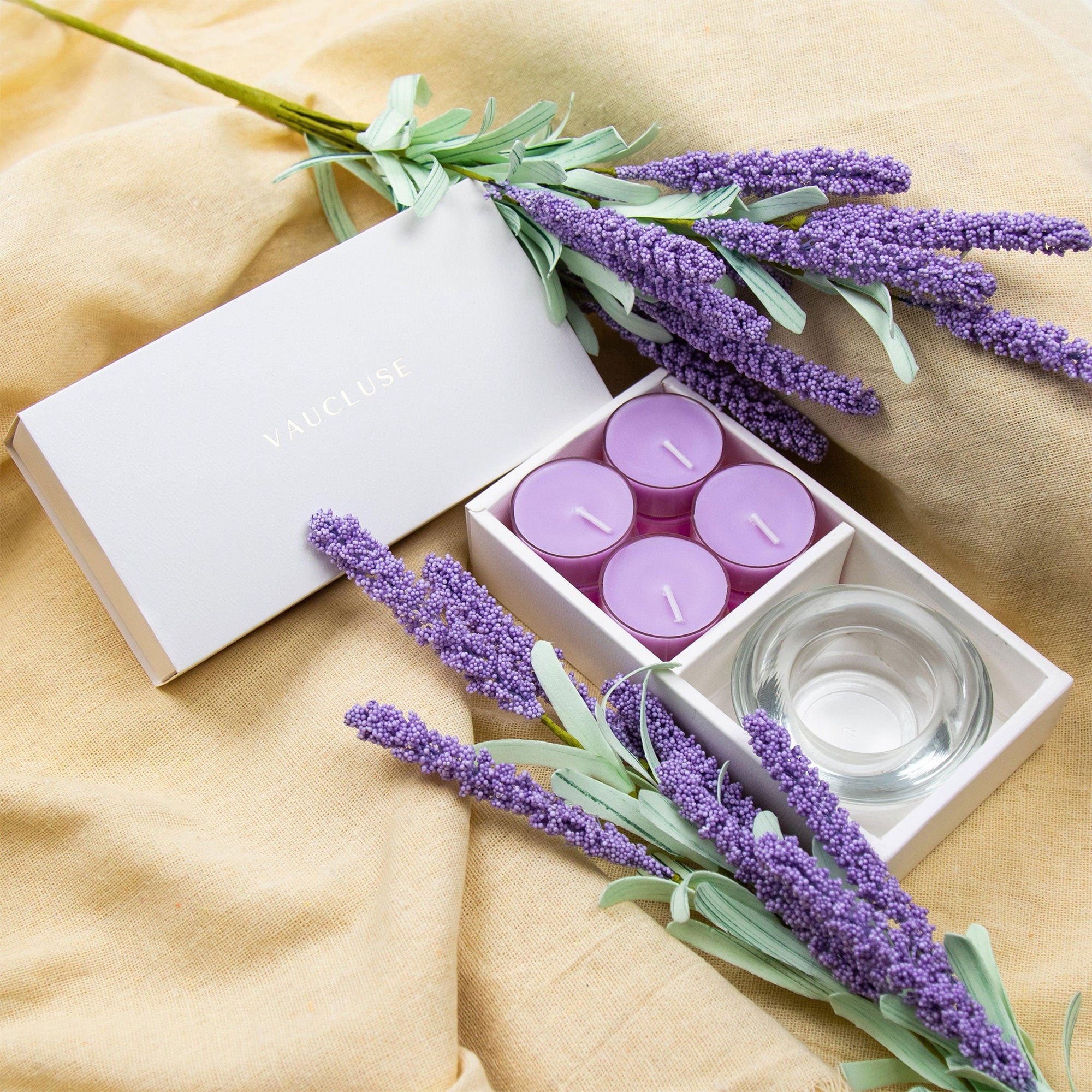Cozy aromatherapy lifestyle scene featuring the open Vaucluse lavender tealight set resting on draped beige linen fabric with sprigs of lavender.