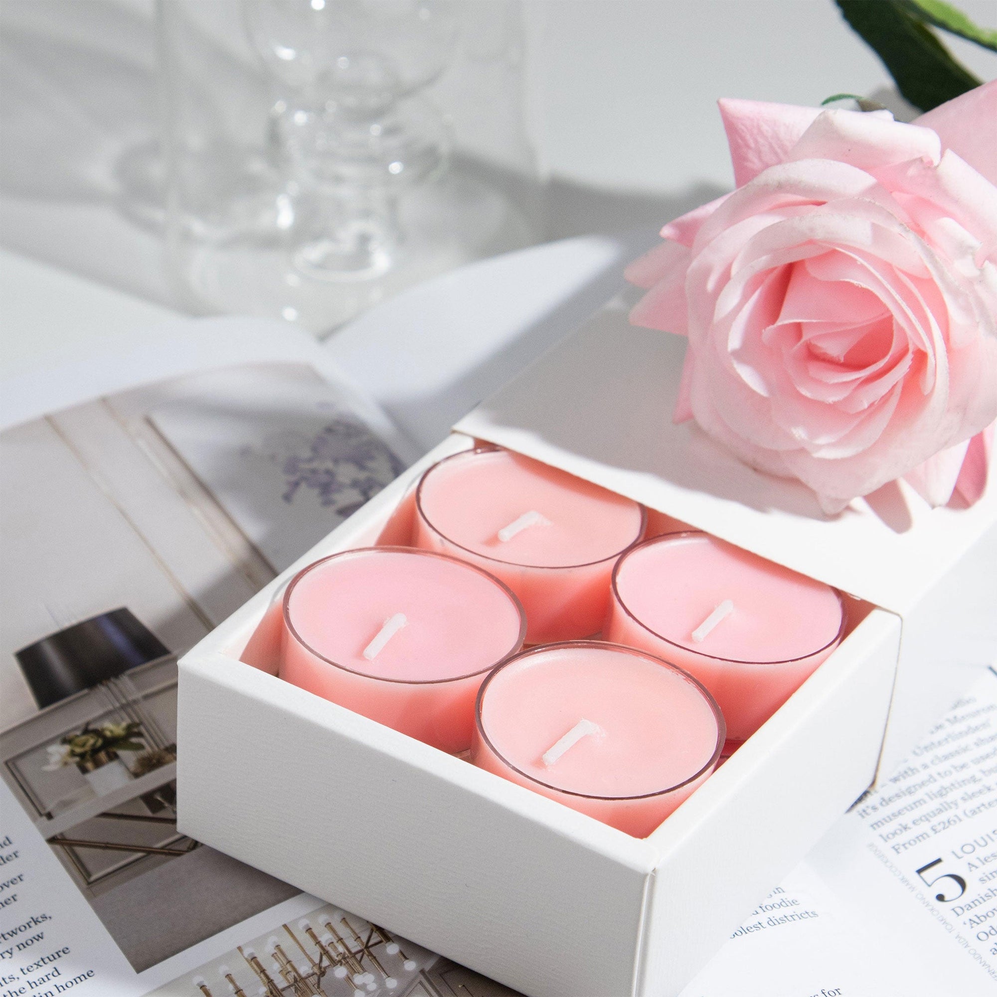 Close-up view of the premium white Vaucluse sliding packaging revealing a tiered 16-pack tray of pink rose scented tealight candles next to an elegant rose blossom.