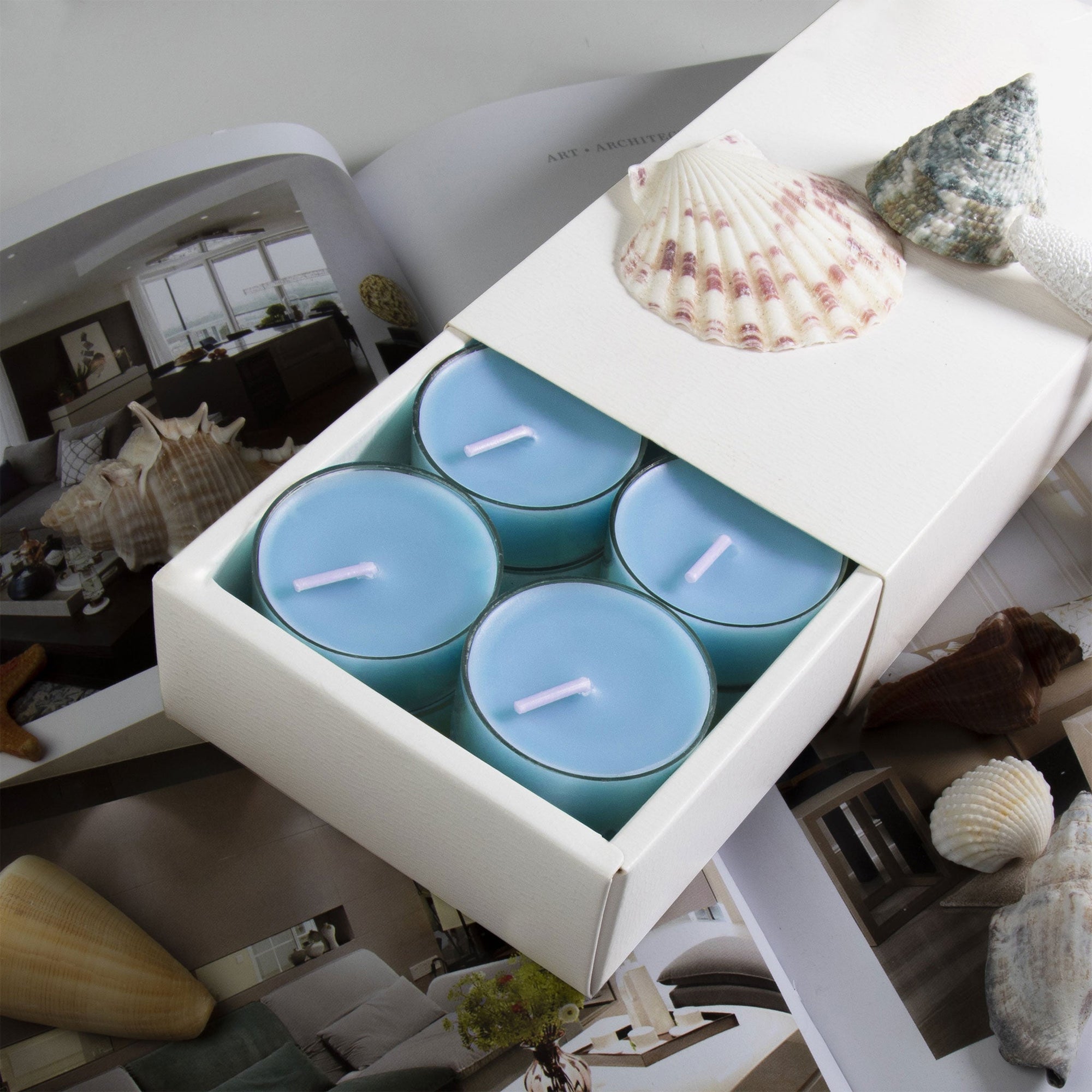A premium white Vaucluse 16-pack box sliding open to reveal a tier of light blue sea breeze tealight candles, decorated with a textured white seashell.