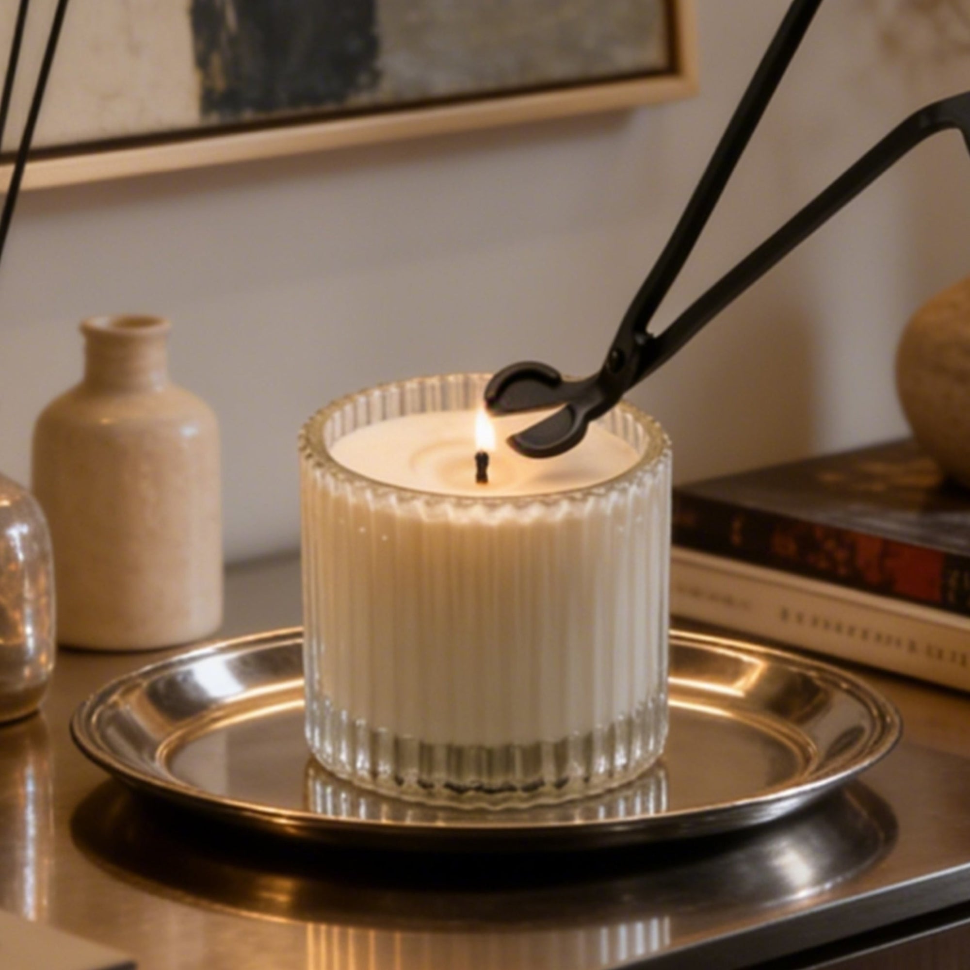 Trimming the wick of a lit luxury white candle in a ribbed glass jar using a matte black candle wick trimmer.