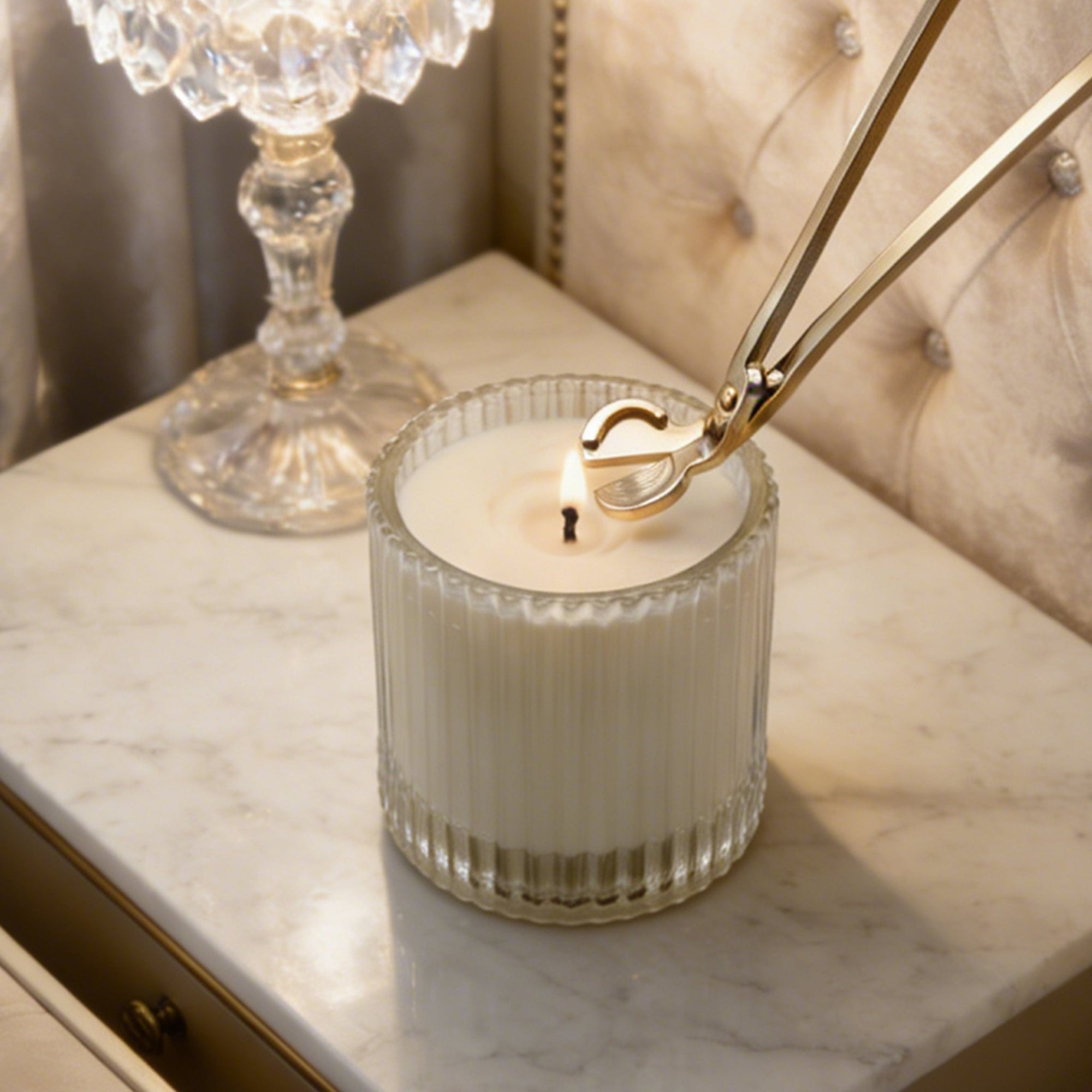 Trimming a candle wick with a polished silver wick trimmer tool on a lit white candle in a ribbed glass jar.