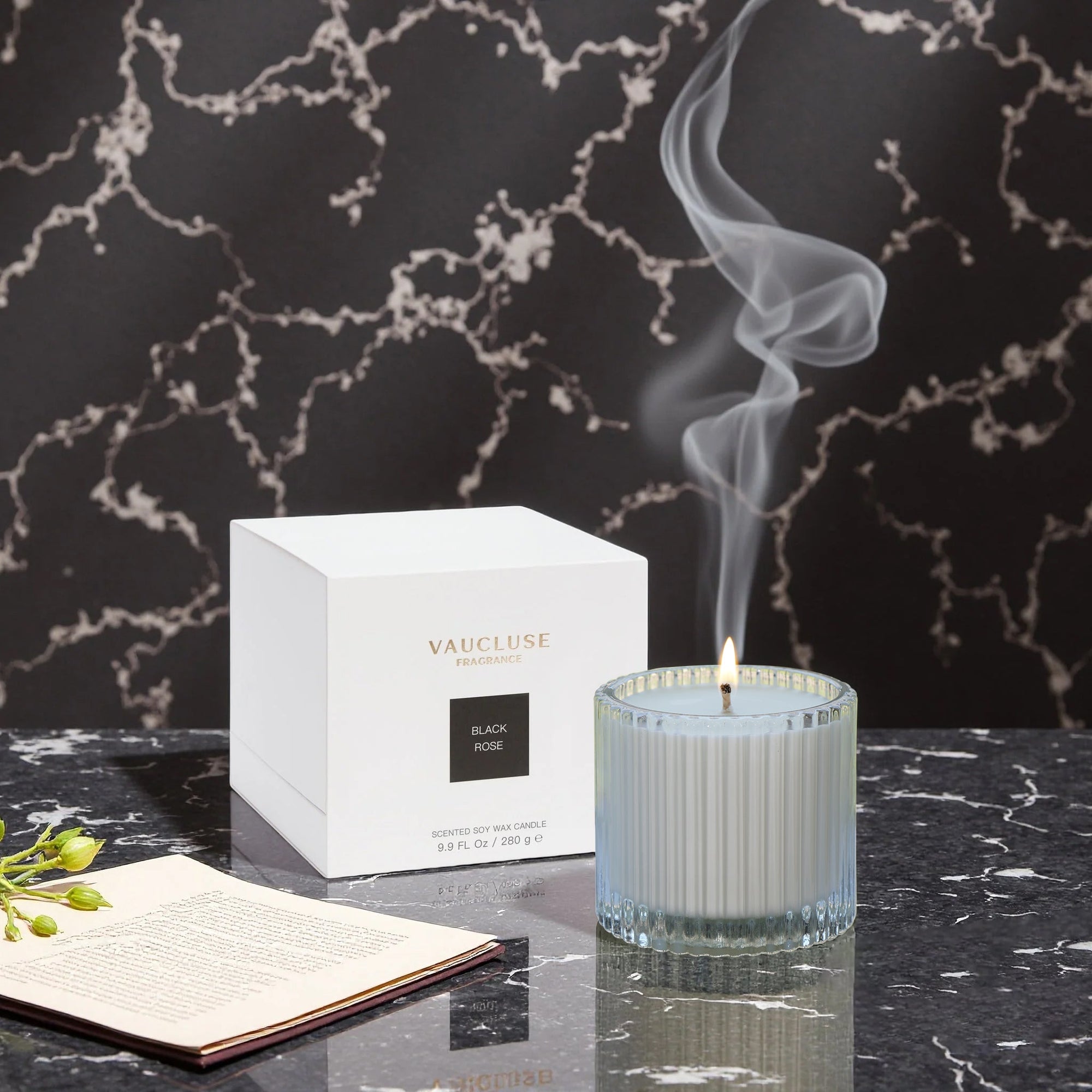 A lit Vaucluse Black Rose scented candle emitting a wisp of smoke, placed next to its minimalist white packaging box on a dark marble surface.