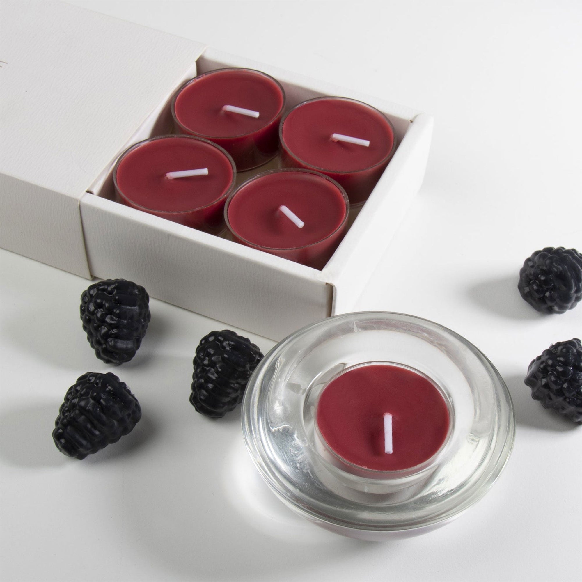 An overhead top-down view of two deep purple blackberry tealight candles in modern clear glass holders resting next to a white embossed Vaucluse gift box.