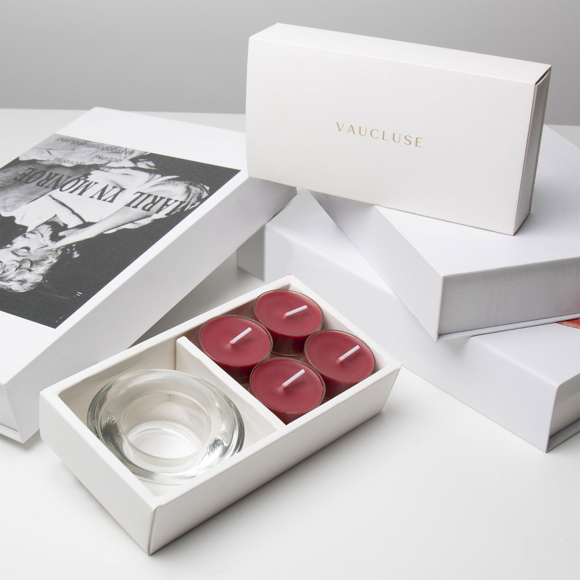 Premium retail unboxing presentation featuring the open burgundy blackberry tealight set resting against stacked white luxury gift boxes with gold branding.