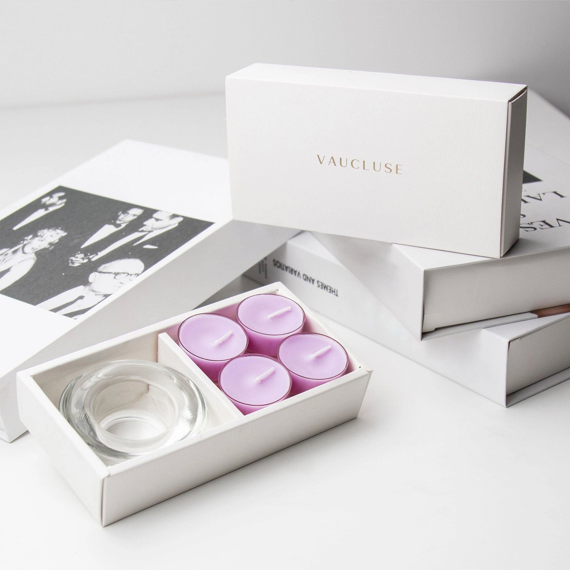 Premium brand unboxing layout showing the open lavender tealight set against a clean backdrop of stacked white luxury gift boxes with gold foil branding.