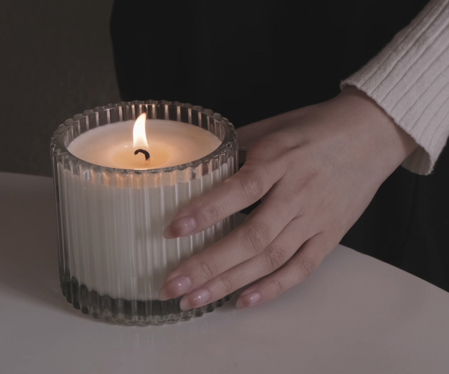 A hand resting next to a lit Vaucluse Fragrance scented candle in a ribbed clear glass jar on a white table.
