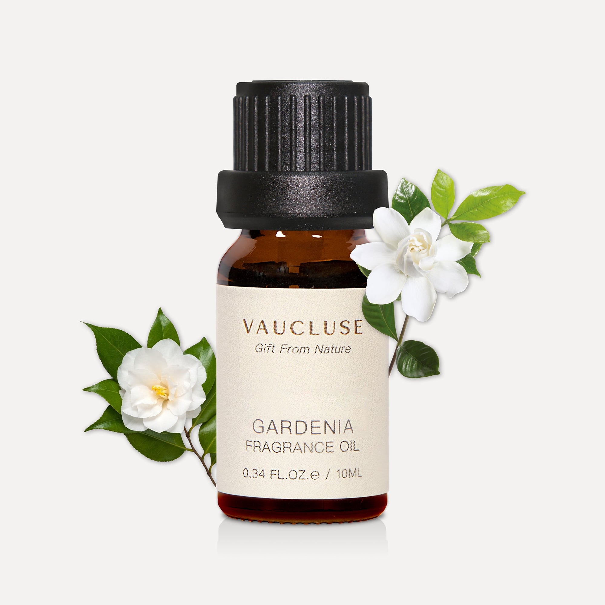A 10ml amber glass bottle of Vaucluse Gardenia Essential Oil decorated with blooming white gardenia flowers and green leaves.