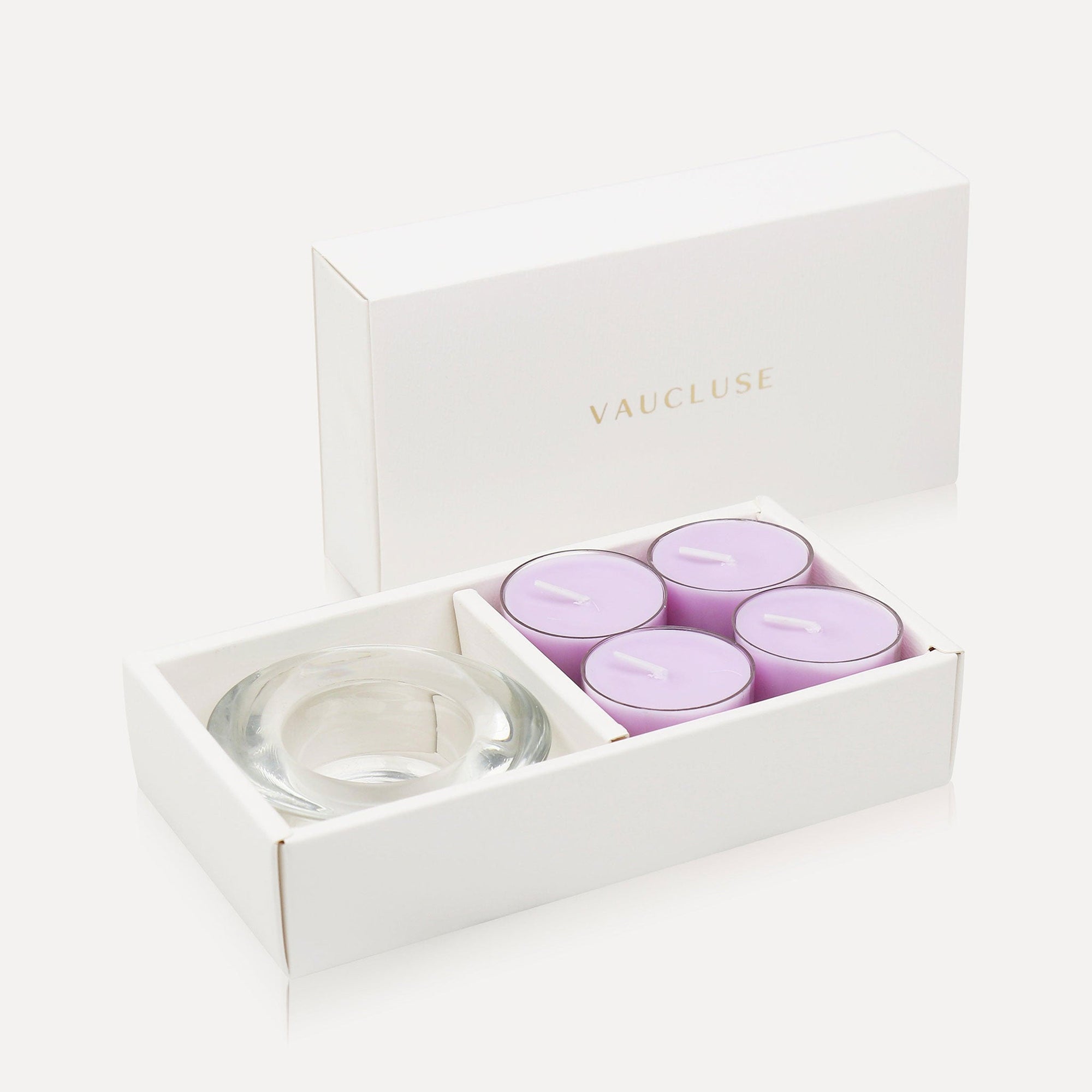 An open luxury white gift box elegantly presenting a set of four light purple lavender tealight candles alongside a thick clear glass candle holder.