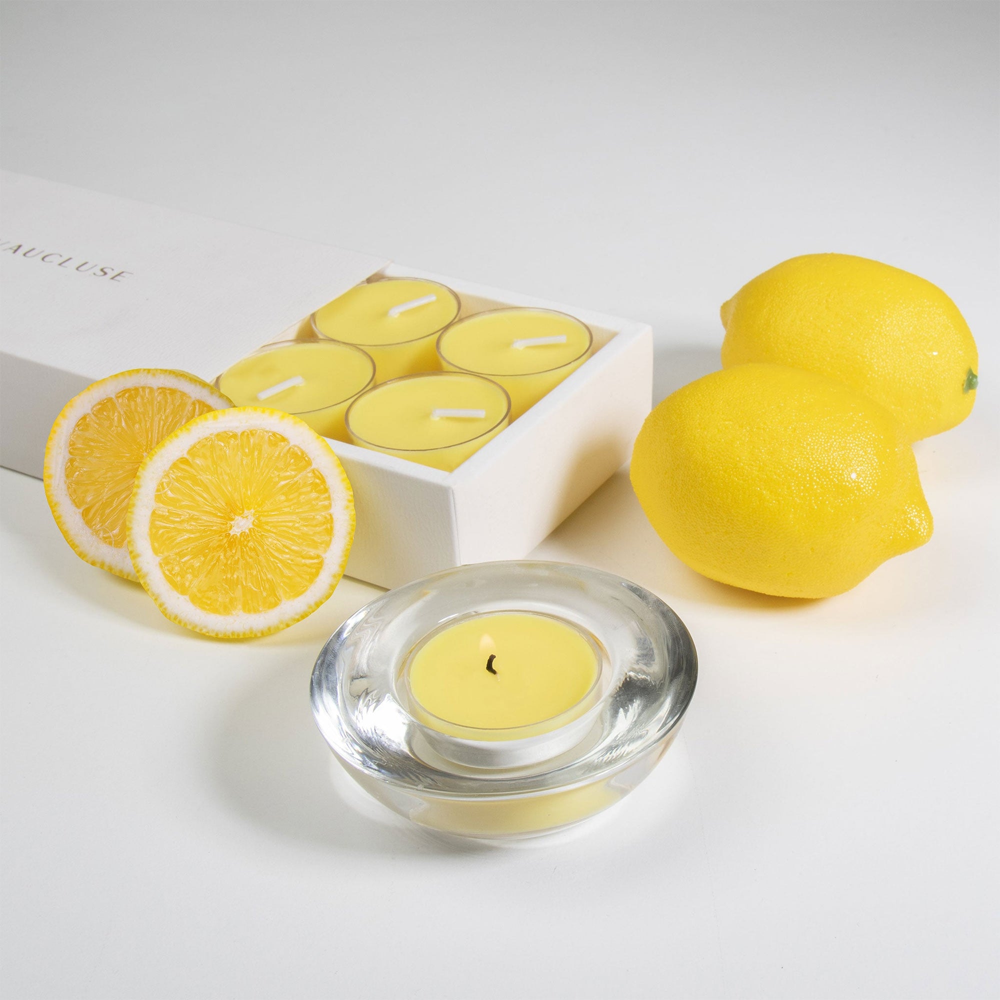 A luxury white Vaucluse 16-pack box sliding open to reveal a tray of yellow lemon tealight candles, shown next to a burning candle in a clear glass holder and whole fresh lemons.