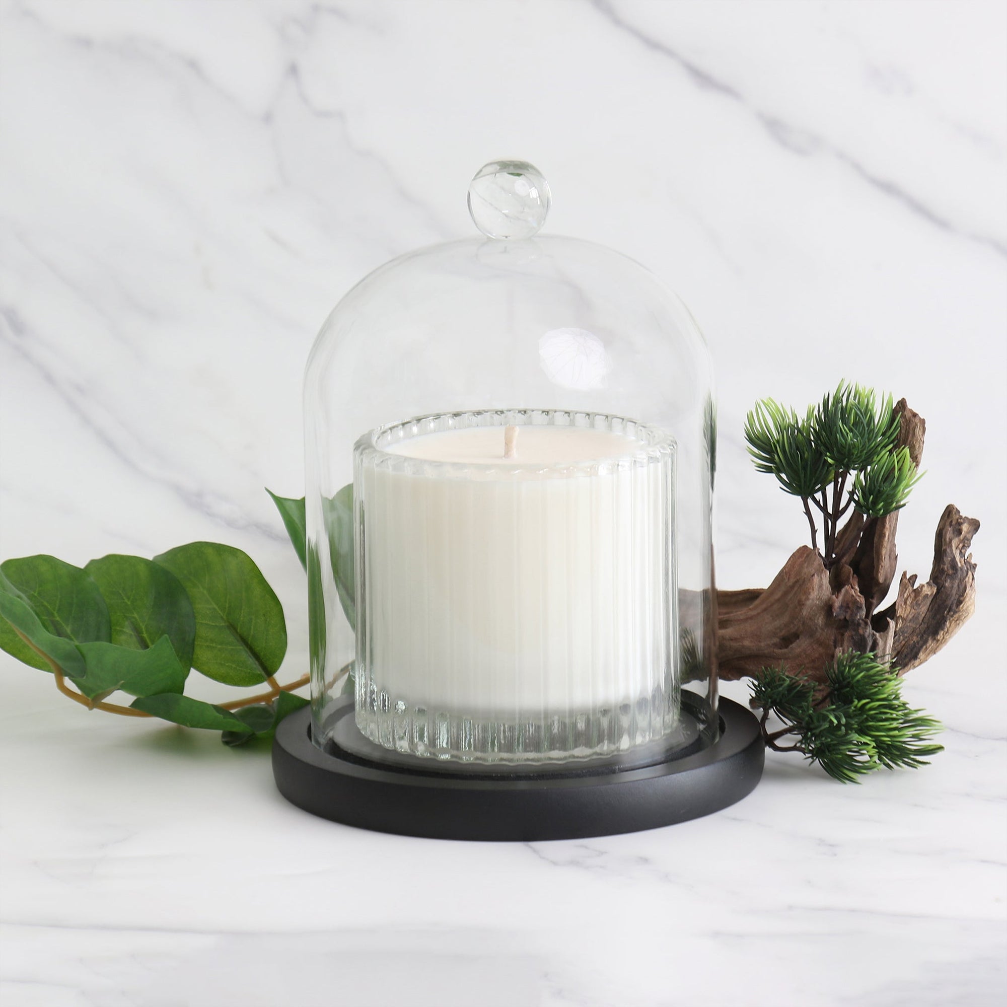 A Vaucluse clear glass dome covering a white scented candle on its black wooden base, styled with fresh green leaves on a bright marble background.