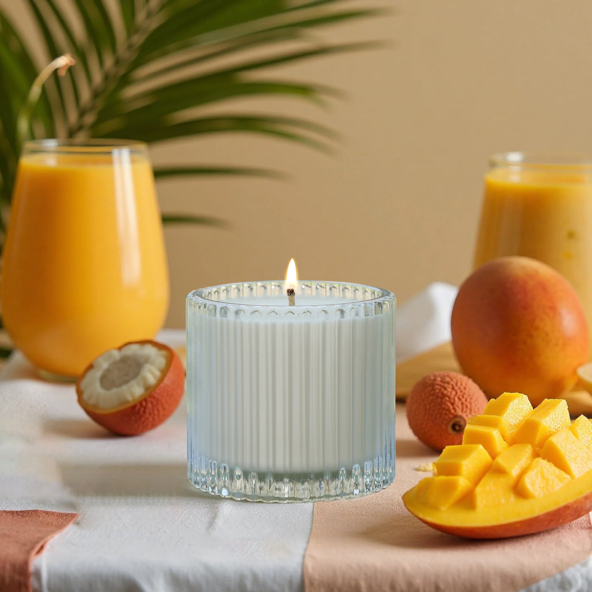 A lit Vaucluse Lychee luxury scented candle glass jar surrounded by fresh sliced mangoes, lychees, and tropical juice glasses.