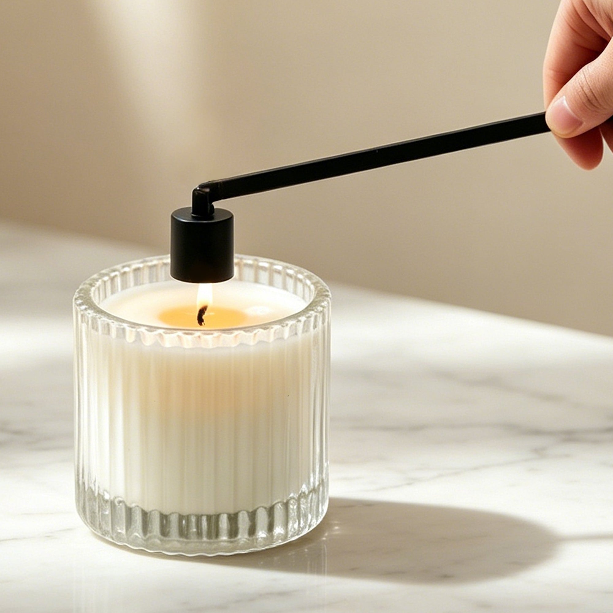 Using a matte black candle snuffer to safely extinguish a burning luxury candle in a ribbed glass jar on a marble tabletop.