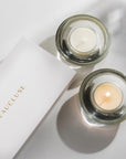 An overhead top-down view of two cream musk tealight candles in modern clear glass holders resting next to a white embossed Vaucluse gift box.