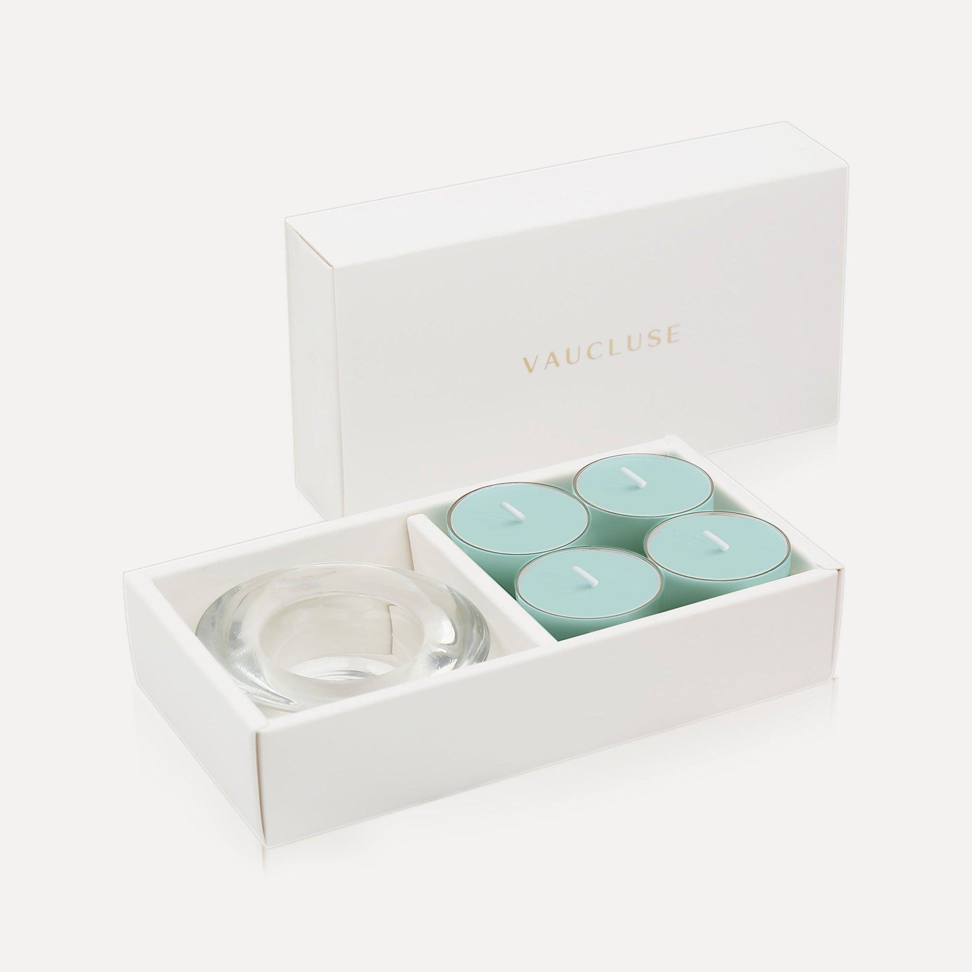 An open white luxury gift box displaying a set of four light aqua-blue ocean tealight candles next to a round clear glass candle holder.