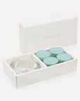 An open white luxury gift box displaying a set of four light aqua-blue ocean tealight candles next to a round clear glass candle holder.