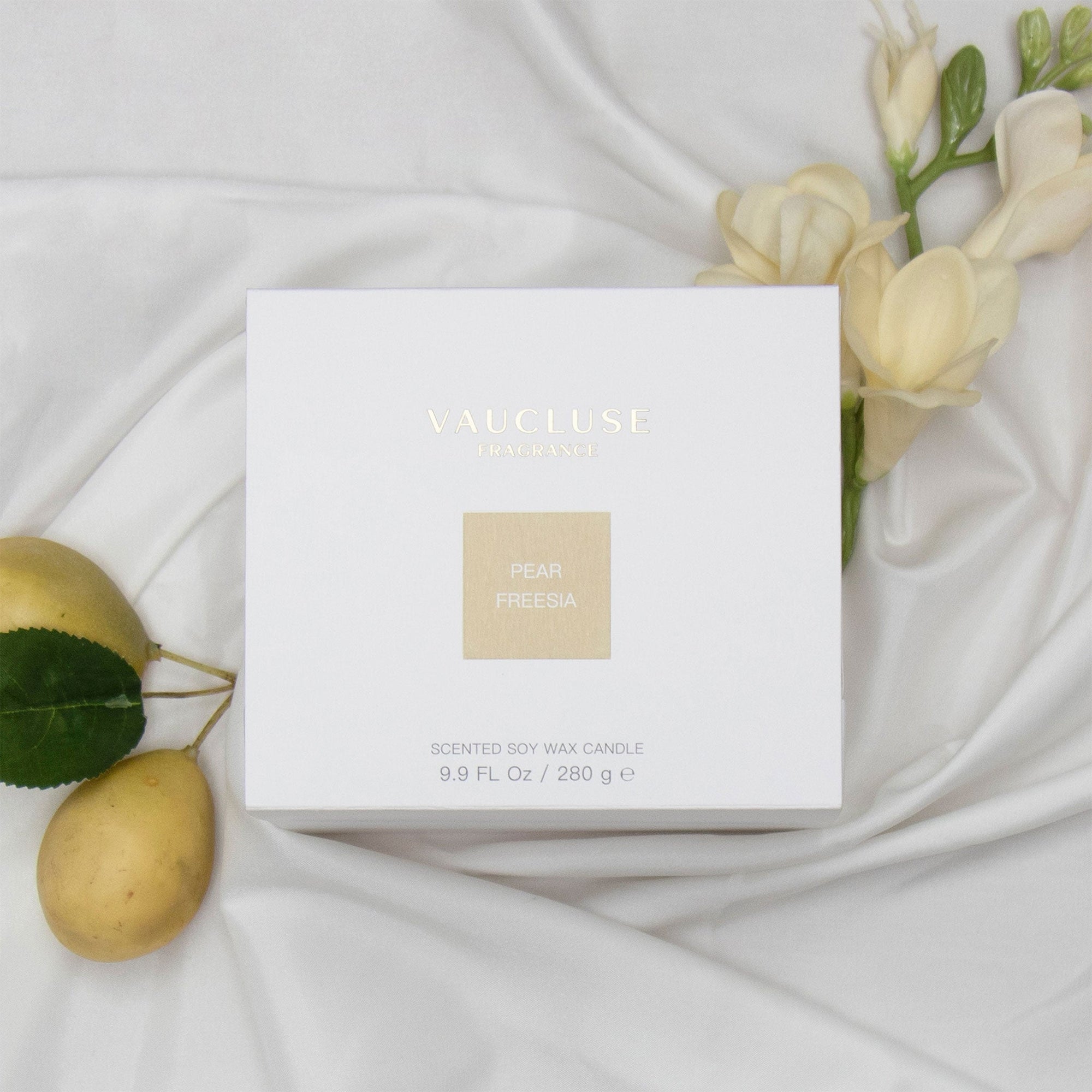 Top view of the minimalist white Vaucluse Pear & Freesia scented wax candle packaging box, decorated with delicate white flowers and fresh pears on a soft silk backdrop.