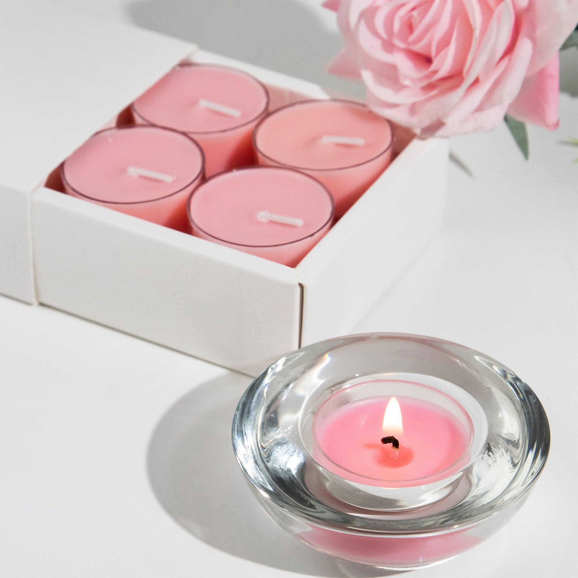 A luxury white Vaucluse 16-pack box sliding open to reveal a tray of pink rose tealight candles, shown next to a burning candle in a clear glass holder and a fresh pink rose.