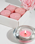A luxury white Vaucluse 16-pack box sliding open to reveal a tray of pink rose tealight candles, shown next to a burning candle in a clear glass holder and a fresh pink rose.