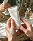 Woman holding Vaucluse Fragrance Rose Velvet hand cream at a vintage vanity table, showing smooth lotion texture on her hand.