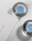 An overhead top-down view of two light blue sea breeze tealight candles resting in modern clear glass holders next to a white embossed Vaucluse gift box.